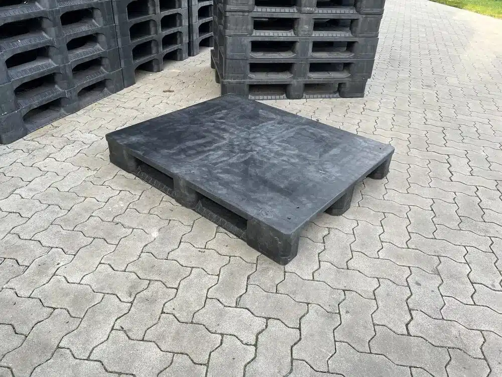 Gebrauchte schwarze Endur i7 Kunststoffpalette mit geschlossenem Deck auf Pflaster, dahinter Palettenstapel