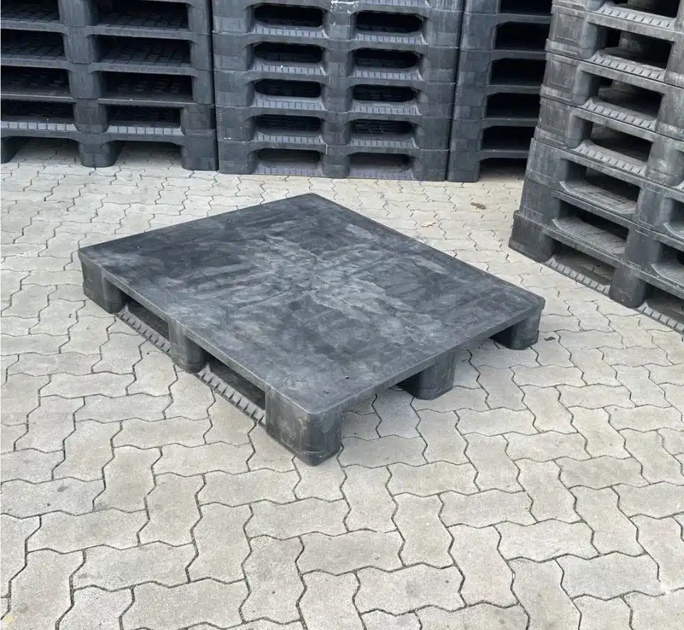 Schwarze Endur i7 CD3R Kunststoffpalette mit geschlossenem Deck liegend auf Pflaster, Palettenstapel im Hintergrund
