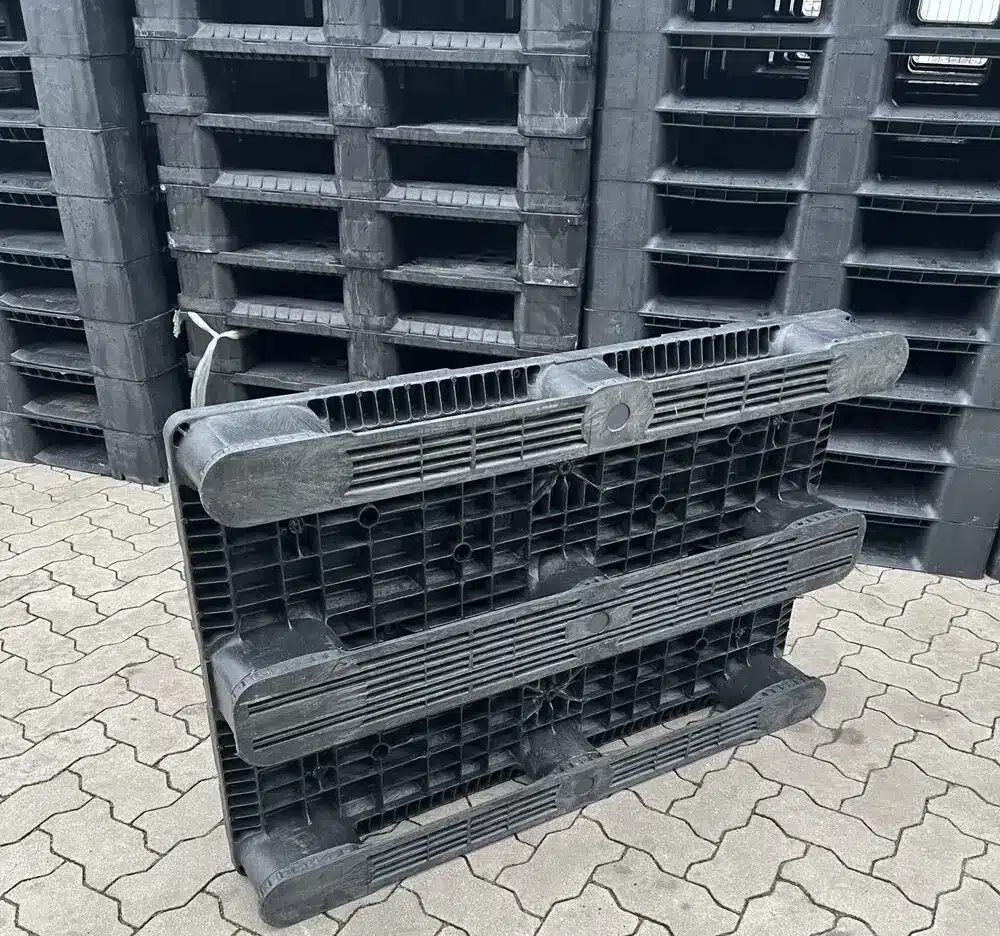 Aufrecht angelehnte Endur i7 Kunststoffpalette, Unterseite mit drei Kufen sichtbar, vor gestapelten Paletten