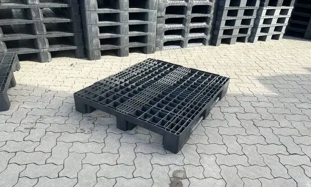 Gebrauchte schwarze Eco E7 Kunststoffpalette mit Gitterdeck auf Pflaster, Palettenstapel im Hintergrund