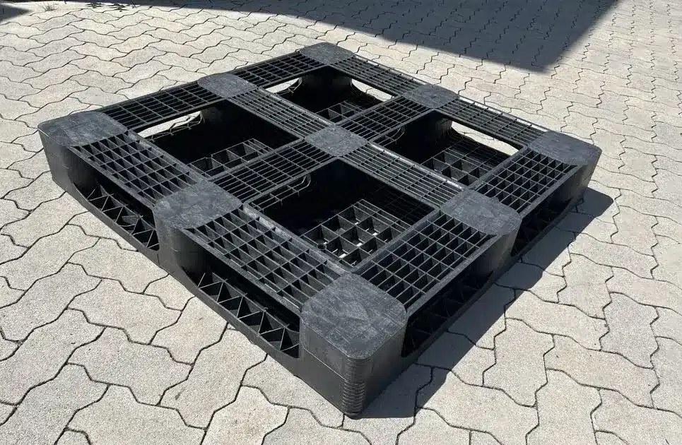 Gebrauchte schwarze CP-9-Kunststoffpalette 1140 x 1140 mm mit offener Zellenstruktur, Schrägansicht auf sonnigem Pflaster