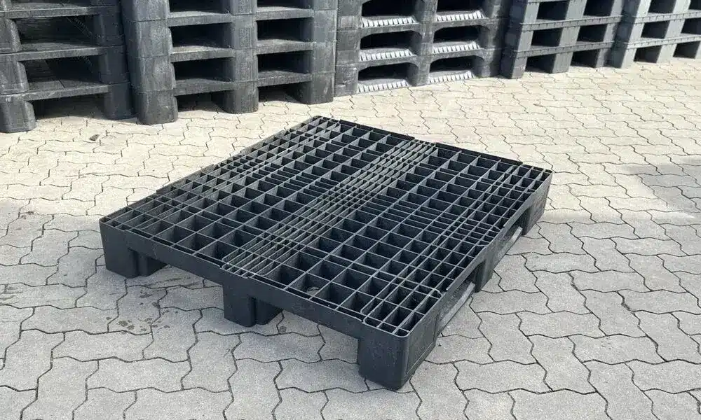 Gebrauchte schwarze Eco E7 Kunststoffpalette, Schrägansicht mit Gitterdeck und drei Kufen auf Pflaster