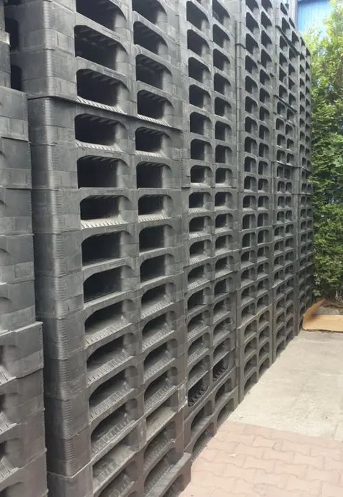 Hoher Stapel gebrauchter schwarzer CP-3-Kunststoffpaletten 1140 x 1140 mm im Außenlager neben grüner Hecke