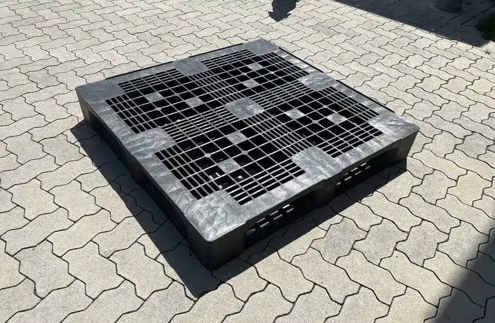 Gebrauchte schwarze CP-9-Kunststoffpalette 1140 x 1140 mm mit feinem Gitterdeck auf Pflaster im Sonnenlicht