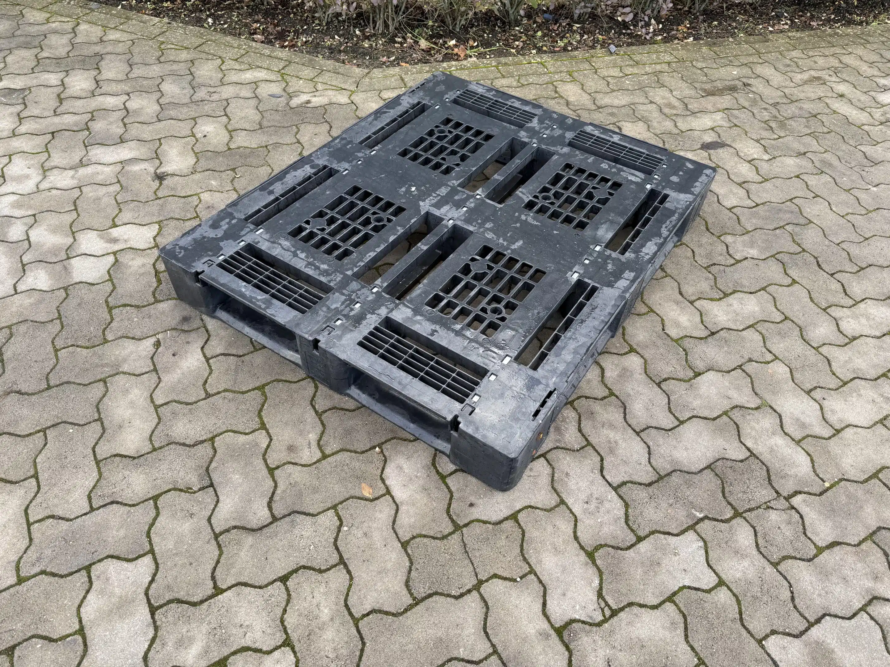 Gebrauchte schwarze Kunststoffpalette mit Gitterstruktur steht auf gepflastertem Boden im Freien.