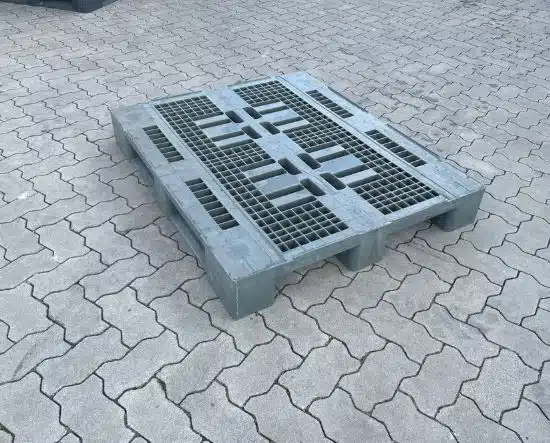 Graue Kunststoffpalette mit Gitteroberdeck auf gepflastertem Boden im Freien.